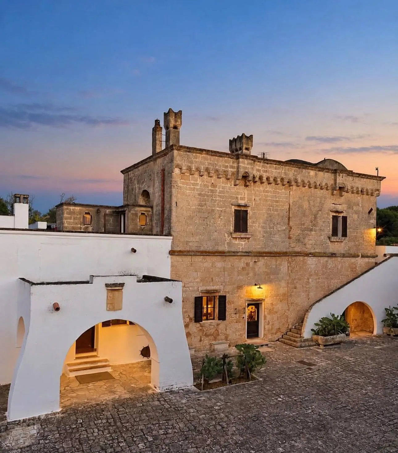 Masseria La Zanzara at dusk, a honey-coloured stone tower and white arched courtyard lit by warm lamps